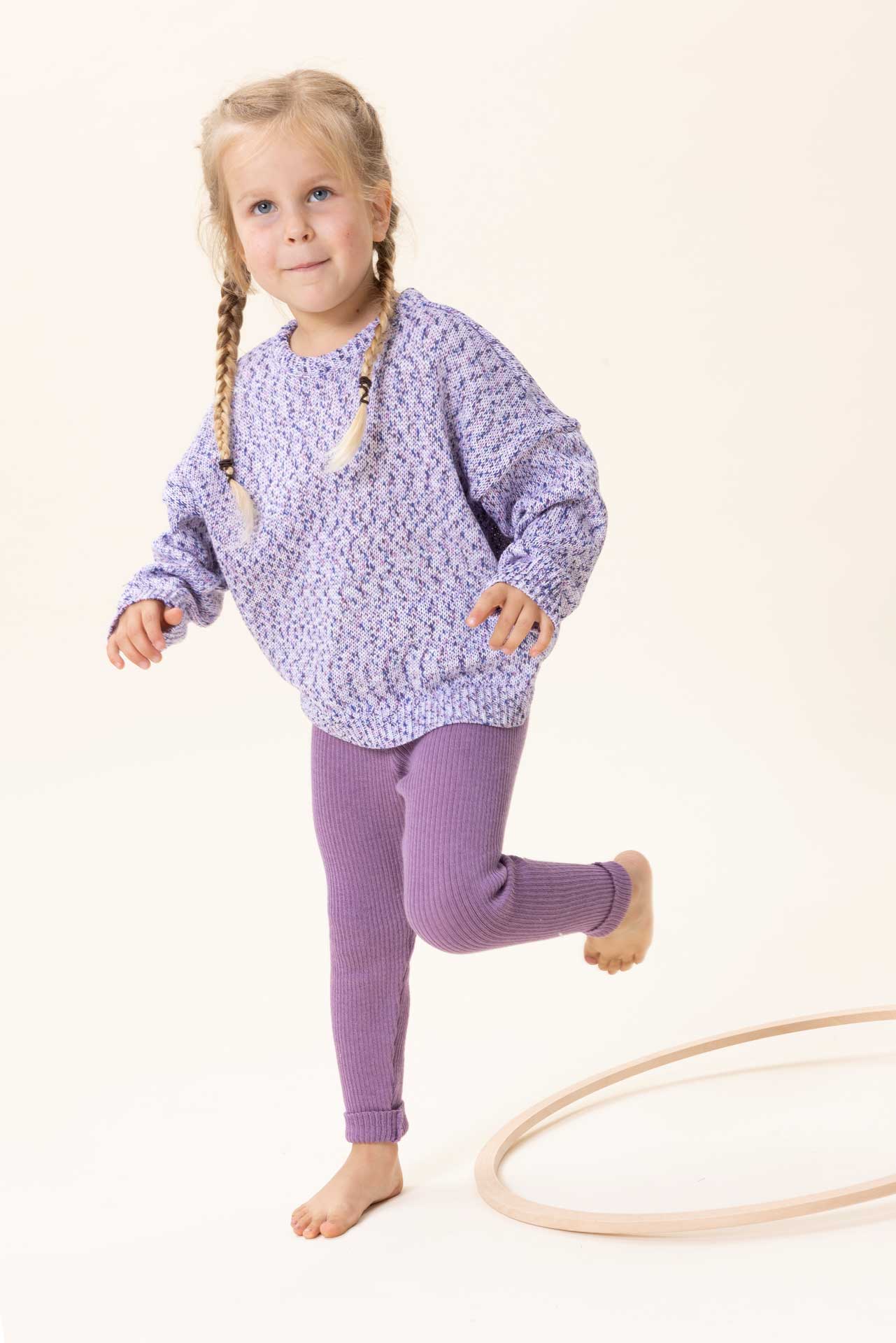 londes Mädchen mit zwei Zöpfen in lila disana Baumwoll-Pullover "Spray" und Leggings spielt mit Hula Hoop Reiden aus Holz"