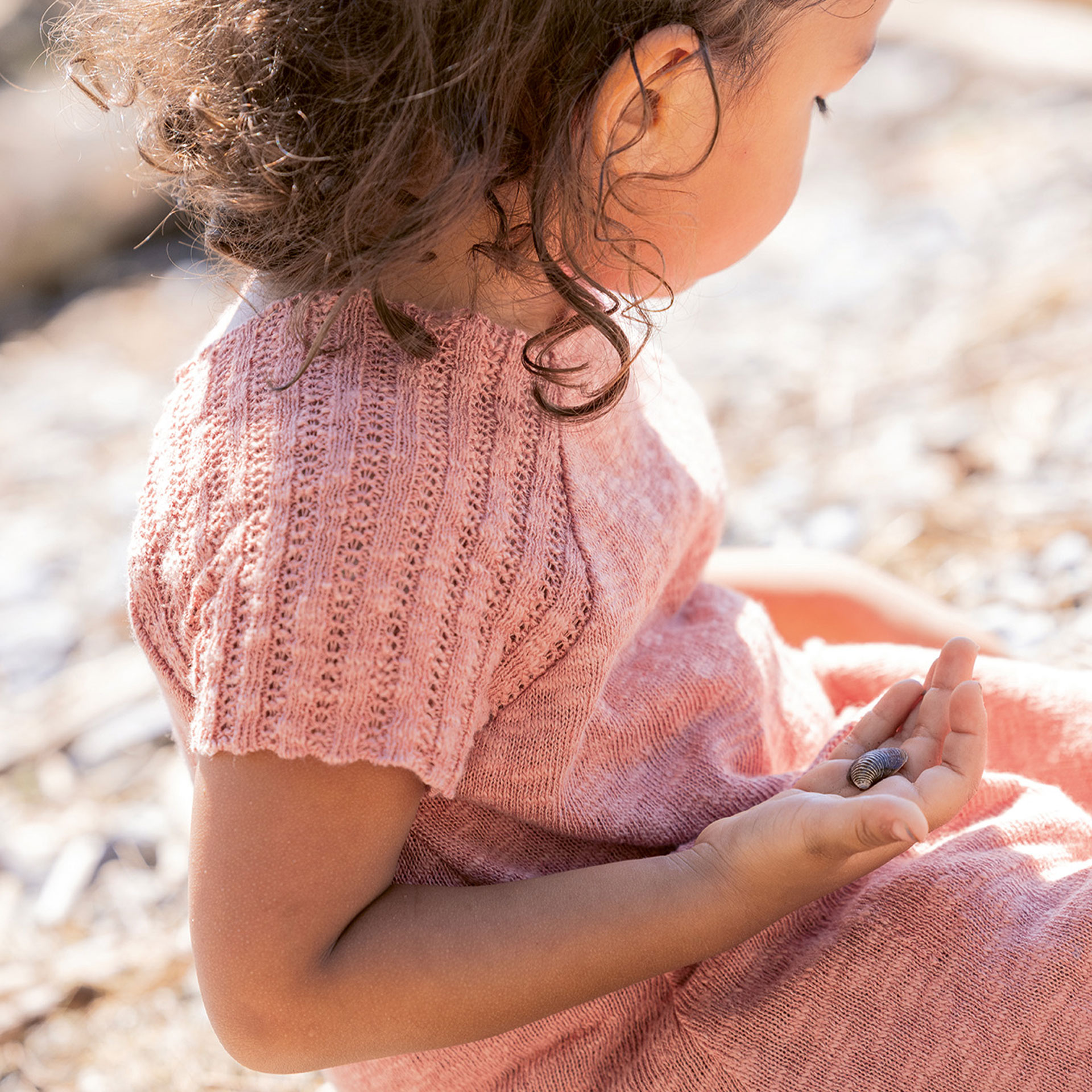 Mädchen mit hellbraunem Hautton in disana Kleid in Softpink sitzt am Kiesstrand und hält eine Muschel in der Hand