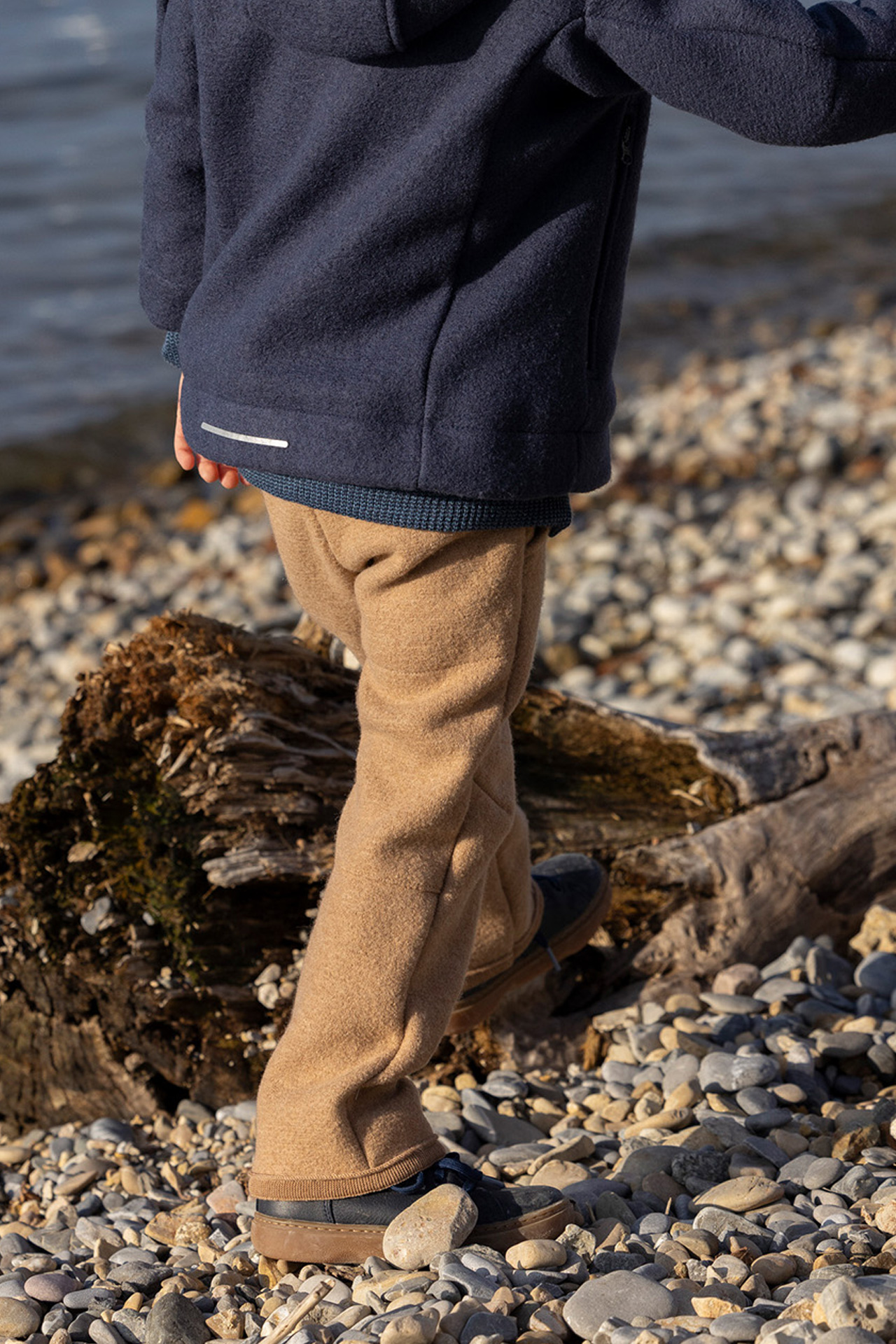 Rückansicht: Kind in indigoblauer disana Outdoor-Jacke & Walk-Hose in Karamell steigt über einen Baumstamm am Kieselstrand