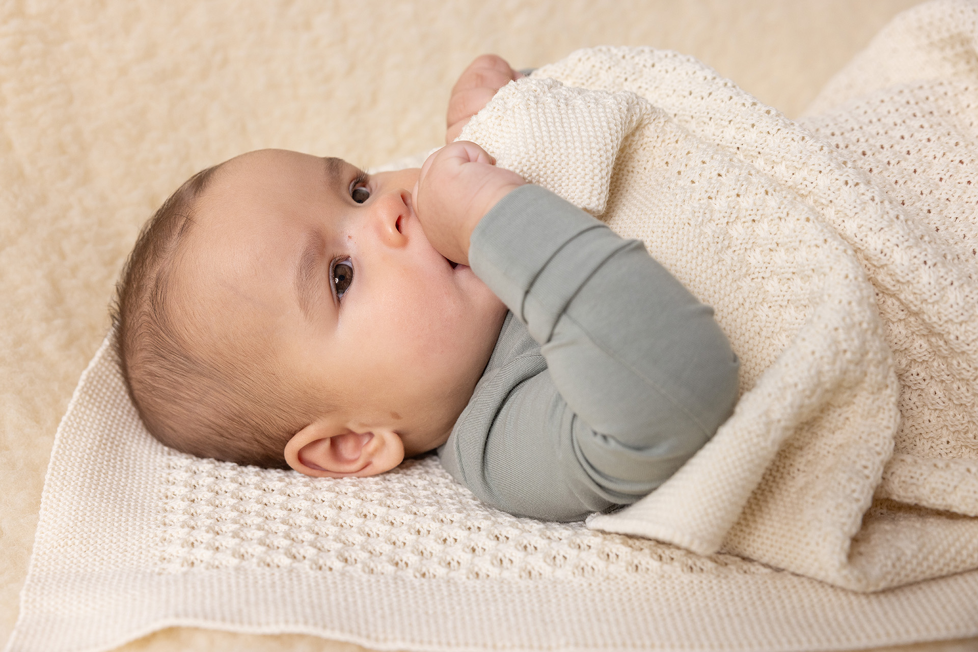 Baby in salbeigrünem Body liegt auf einer cremefarbenen disana Wabenstrick-Decke und zieht sich diese spielerisch zum Mund.