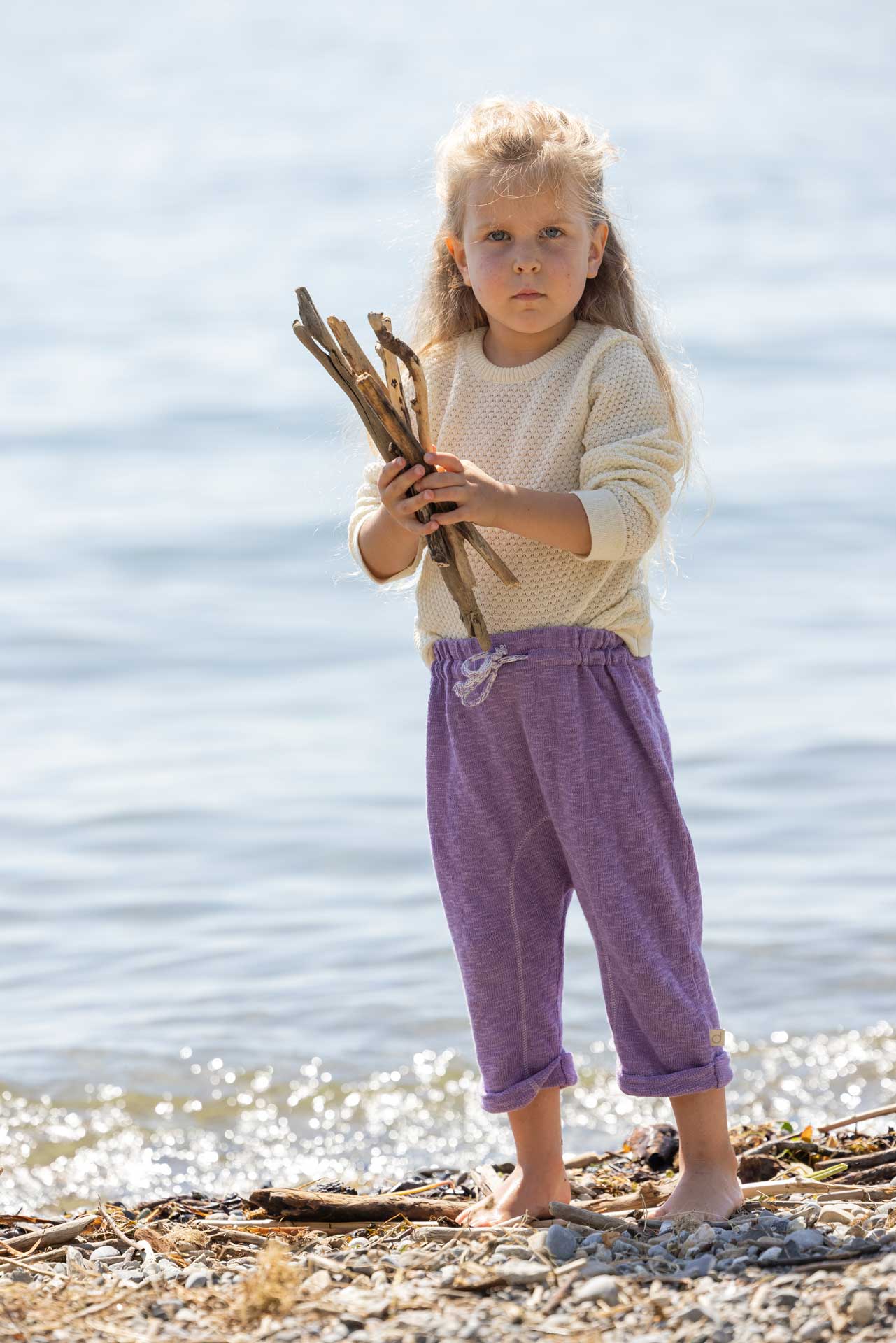 Blondes Mädchen am Seeufer in disana Wabenstrick-Pullover in Natur und Leinen-Strickhose in Flieder hält Treibholz