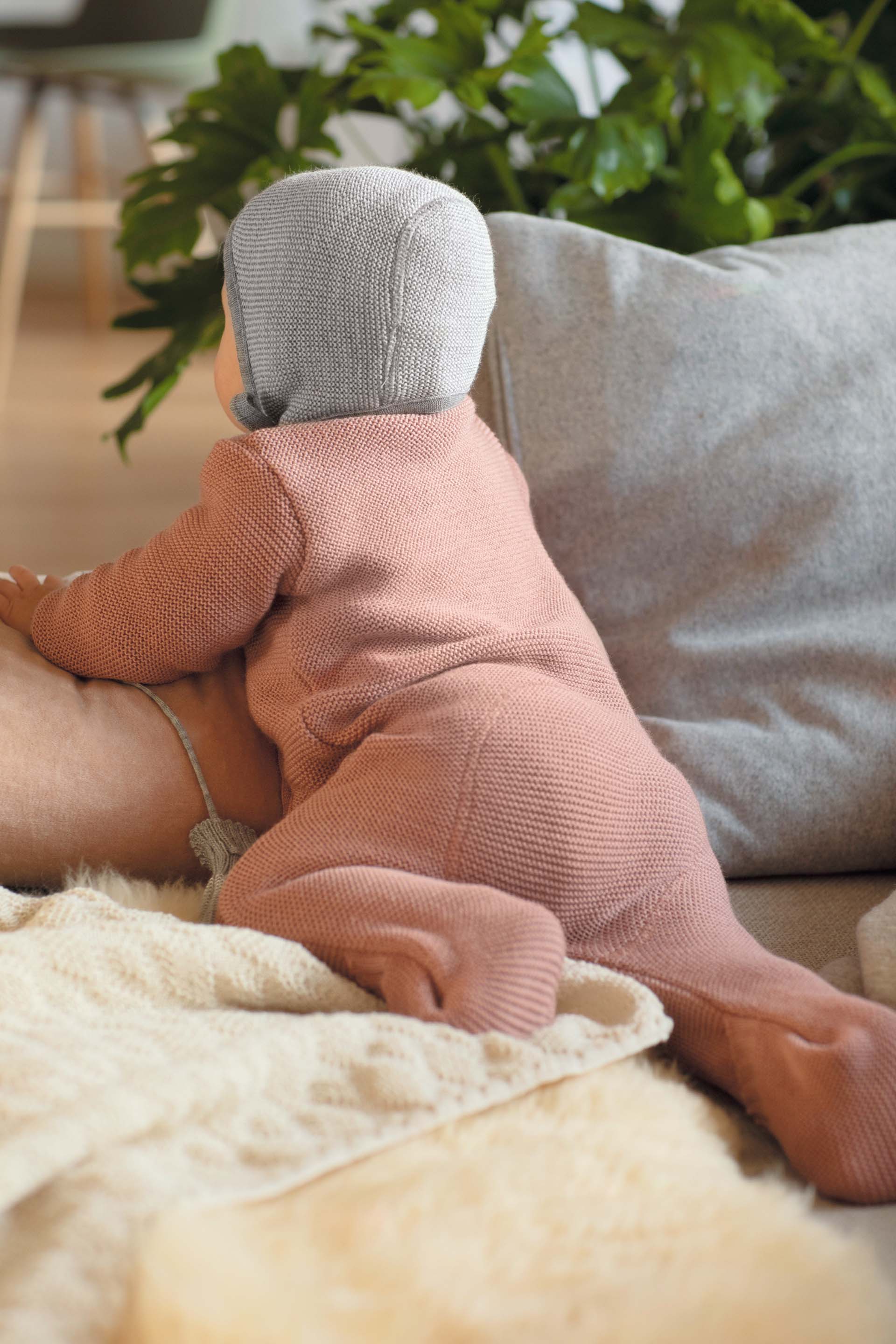 Rückenansicht eines Babys in disana Strick-Overall in Rosé und Strick-Mütze in Grau krabbelt auf einem Lammfell