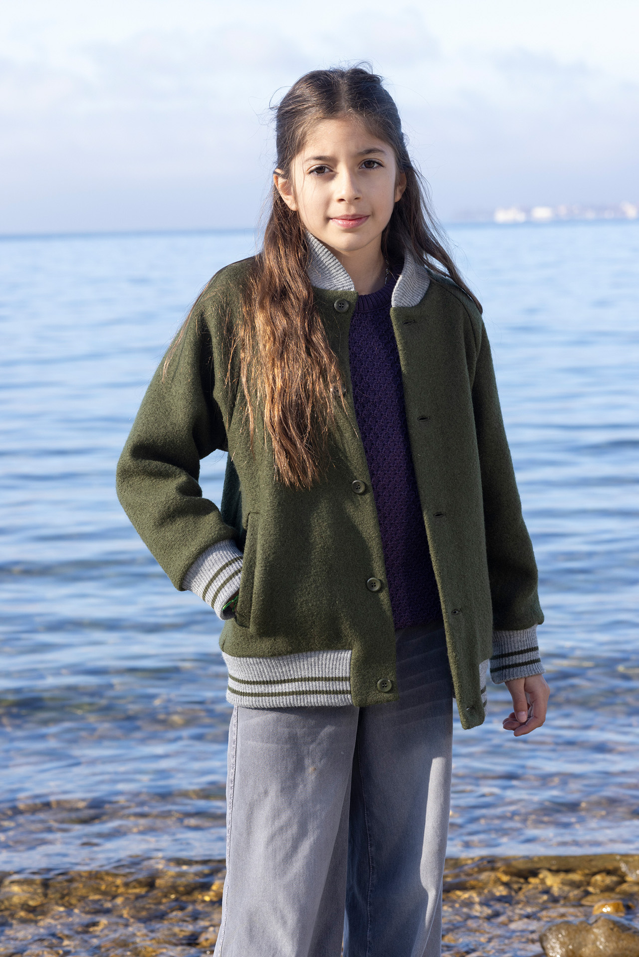 Mädchen mit braunen Haaren in College-Jacke Oliv & Aran-Pullover Aubergine steht am Seeufer, eine Hand in der Tasche
