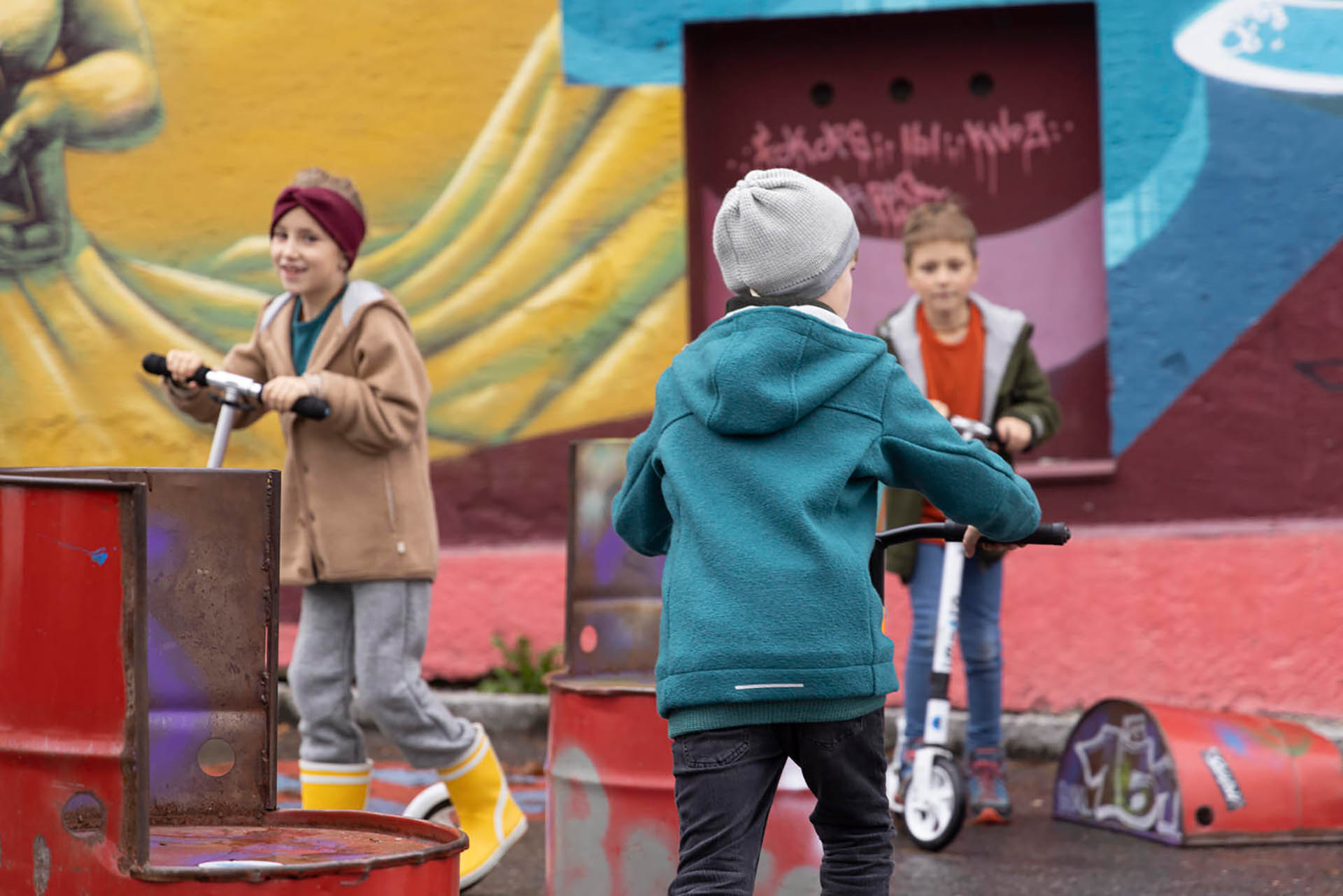 Mehrere Kinder in disana Walk-Kleidung aus Bio-Merinwolle auf Roller vor einer bunten Graffiti-Wand