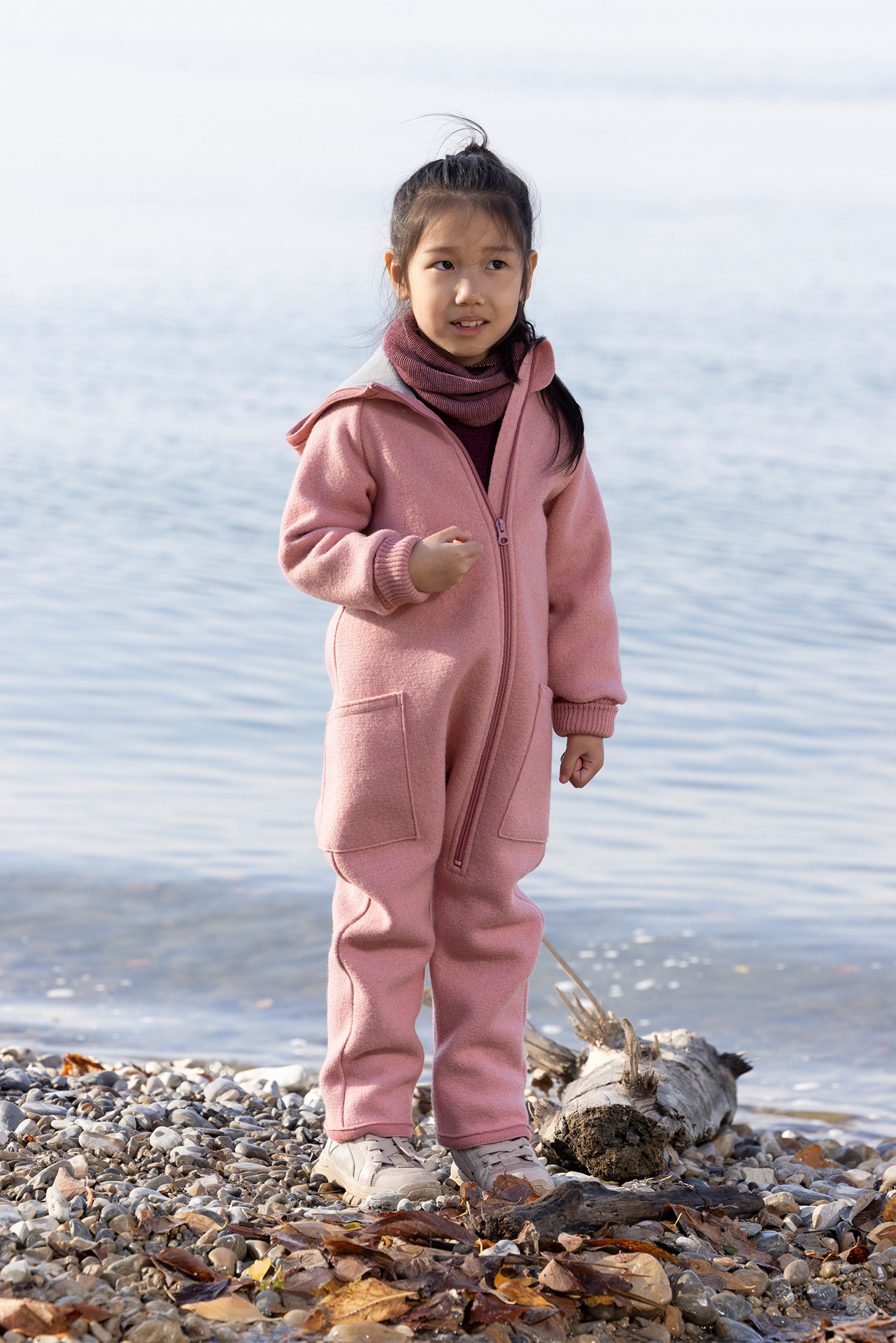 Asiatisches Mädchen in disana Walk-Overall Softpink und Schlauch-Schal in Cassis-Rosé steht am Kieselstrand am Seeufer.