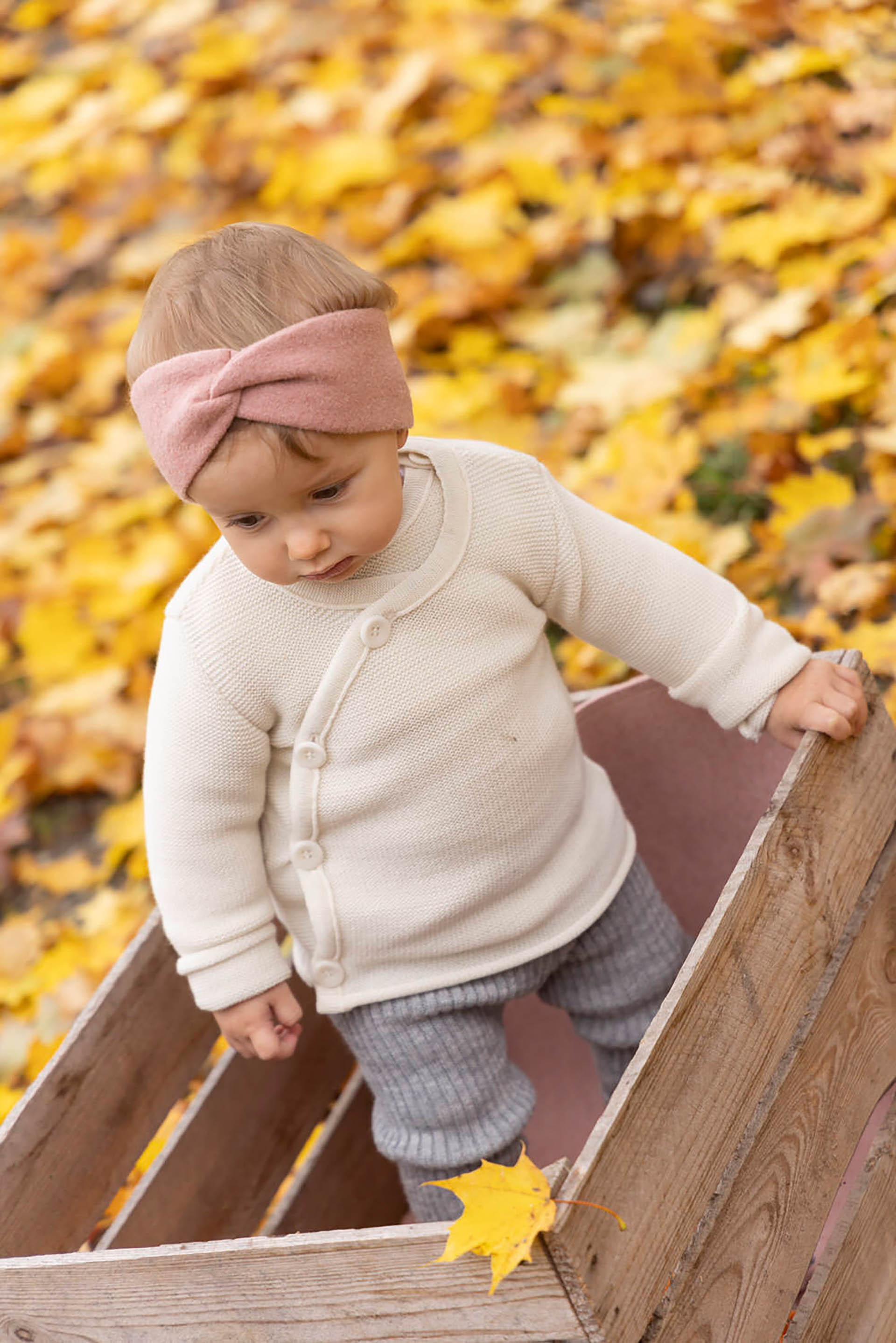 Kleinkind in disana Melange-Jacke Natur Strick-Leggings Grau Stirnband Rosé in einer Holzkiste stehend Hintergrund Herbstlaub