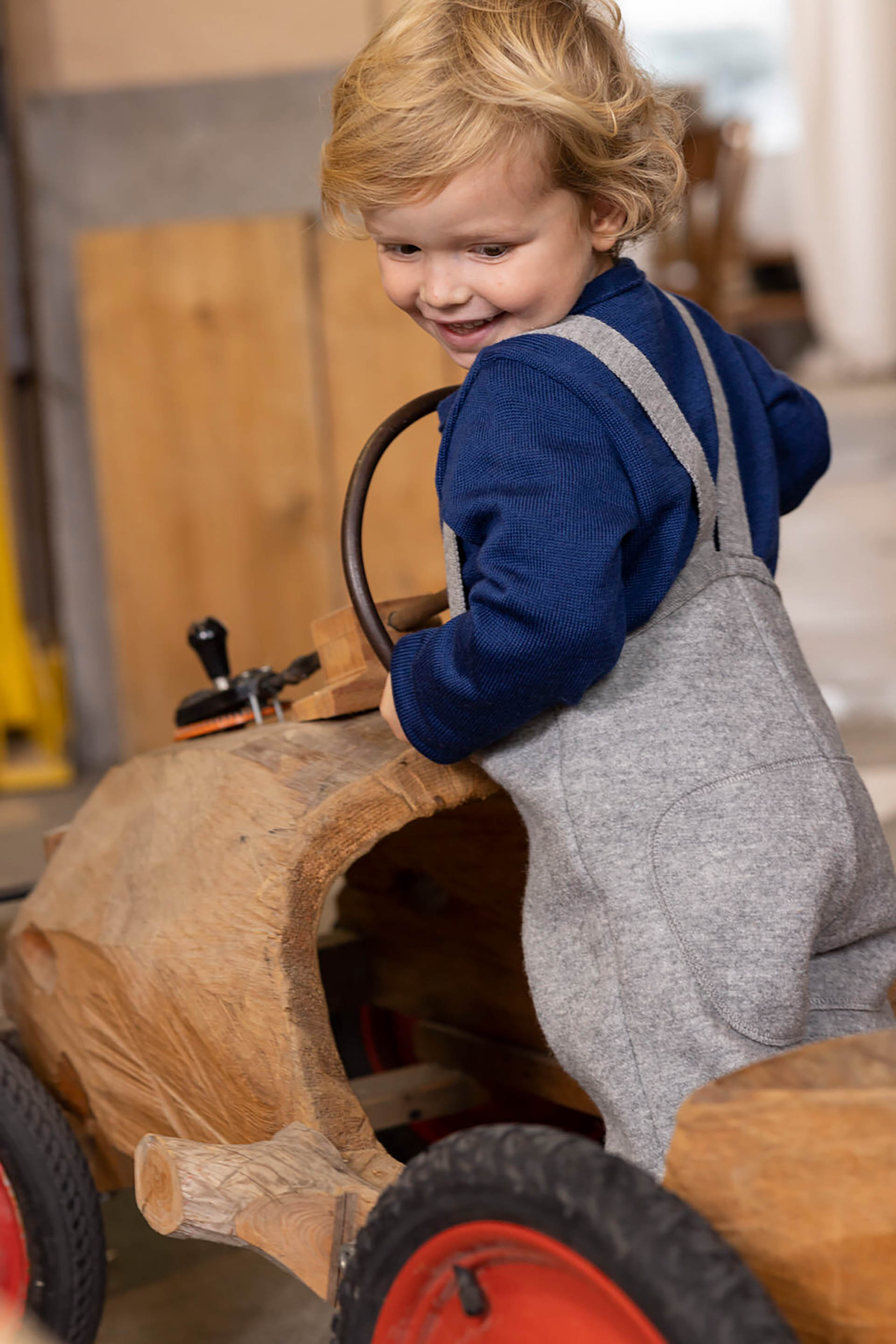 Blondes Kind in disana Walk-Hose mit Trägern in Grau und Strick-Pullover in marine spielt auf einem Holz-Traktor