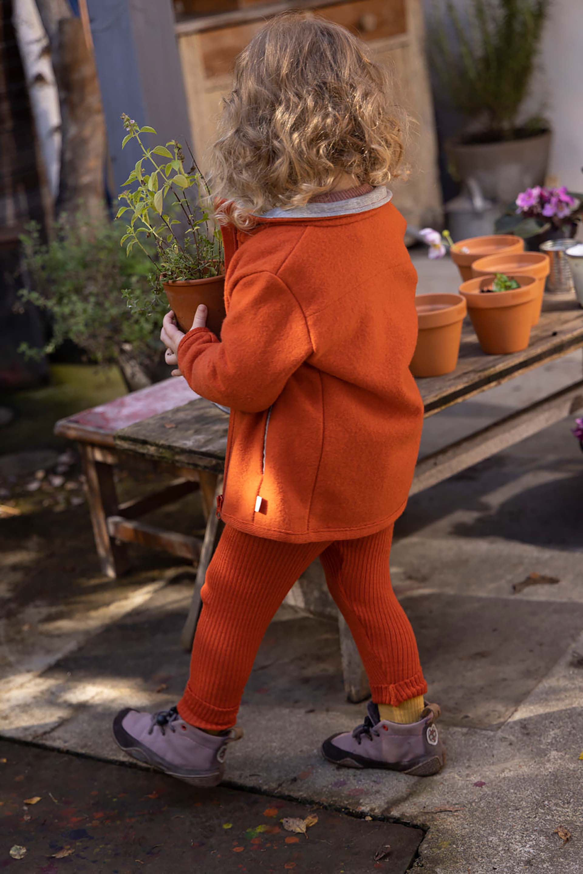 Rückseite eines blonden Kinds mit disana Zippper-Jacke und Leggings in Orange in einem Hof mit Blumentöpfen