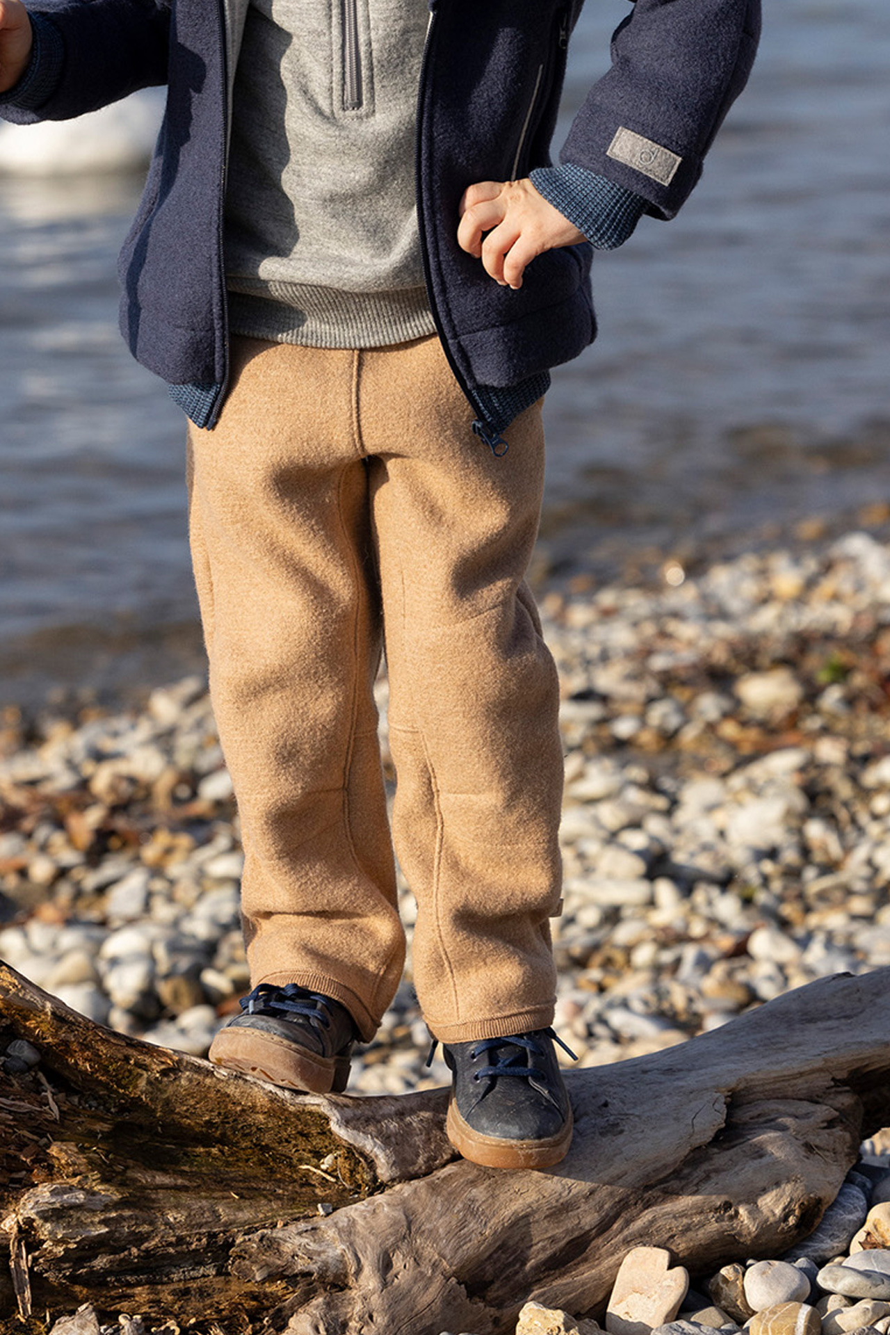 Kind in disana Walk-Hose Karamell & Outdoor-Jacke Indigo steht auf Baumstamm am Seeufer; Fokus auf die Knie-Verstärkungen