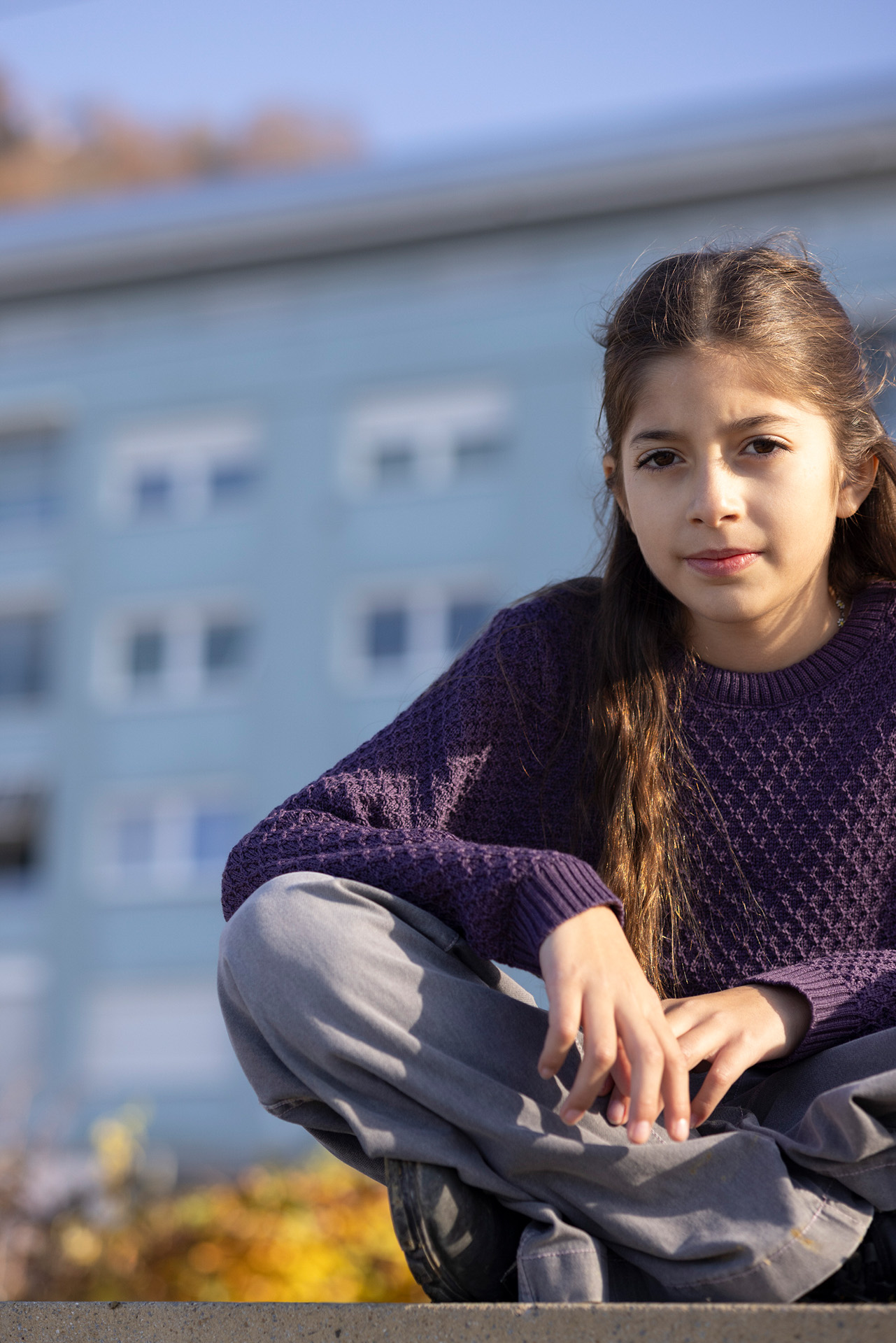 Mädchen, mittelbraune Haaren im disana Aran-Pullover, Aubergine im Schneidersitz vor einem Gebäude im weichen Sonnenlicht