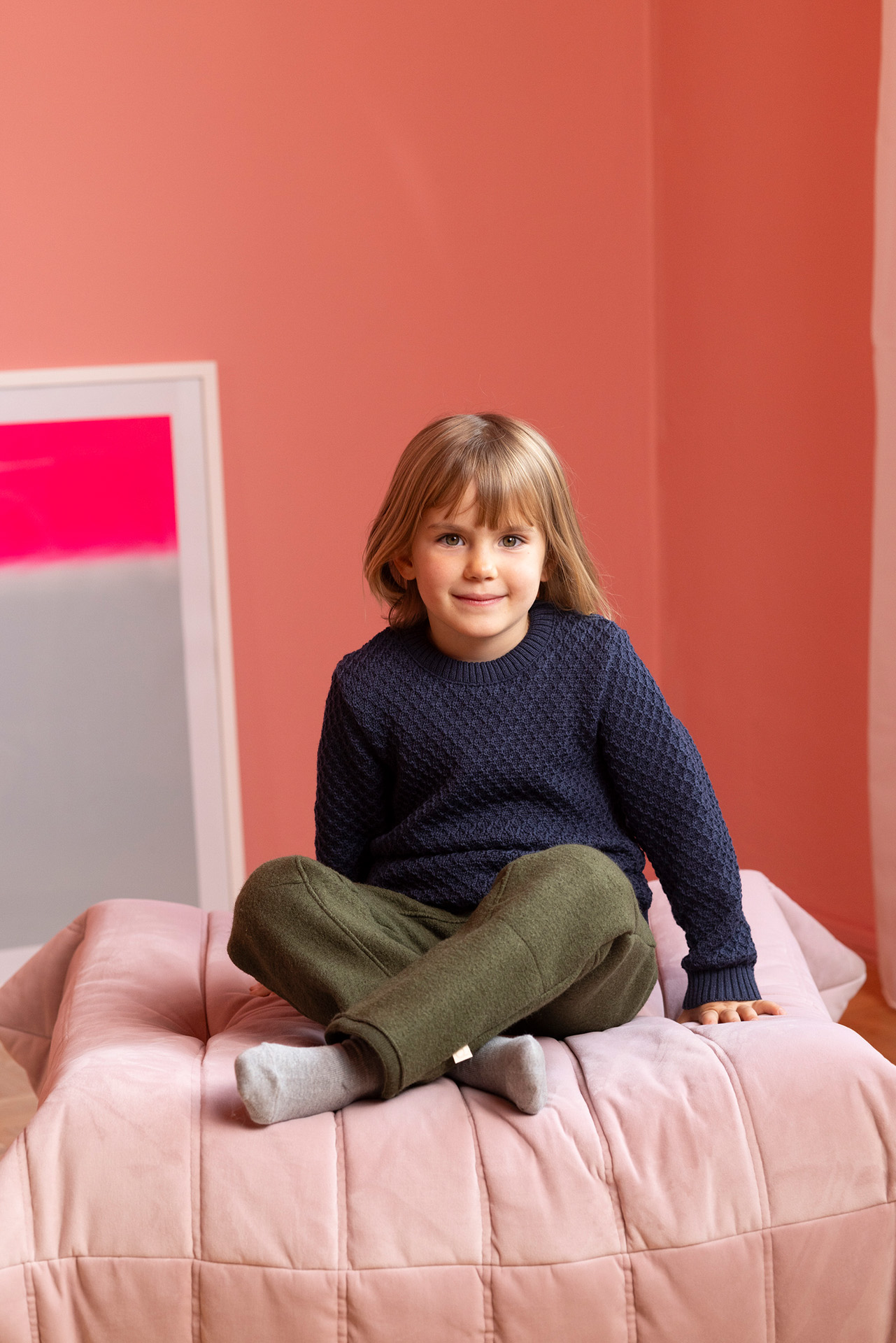 Blondes Kind im disana Aran-Pullover Indigo, Walk-Hose in Oliv sitzt im Schneidersitz auf rosa Polster vor lachsfarbener Wand