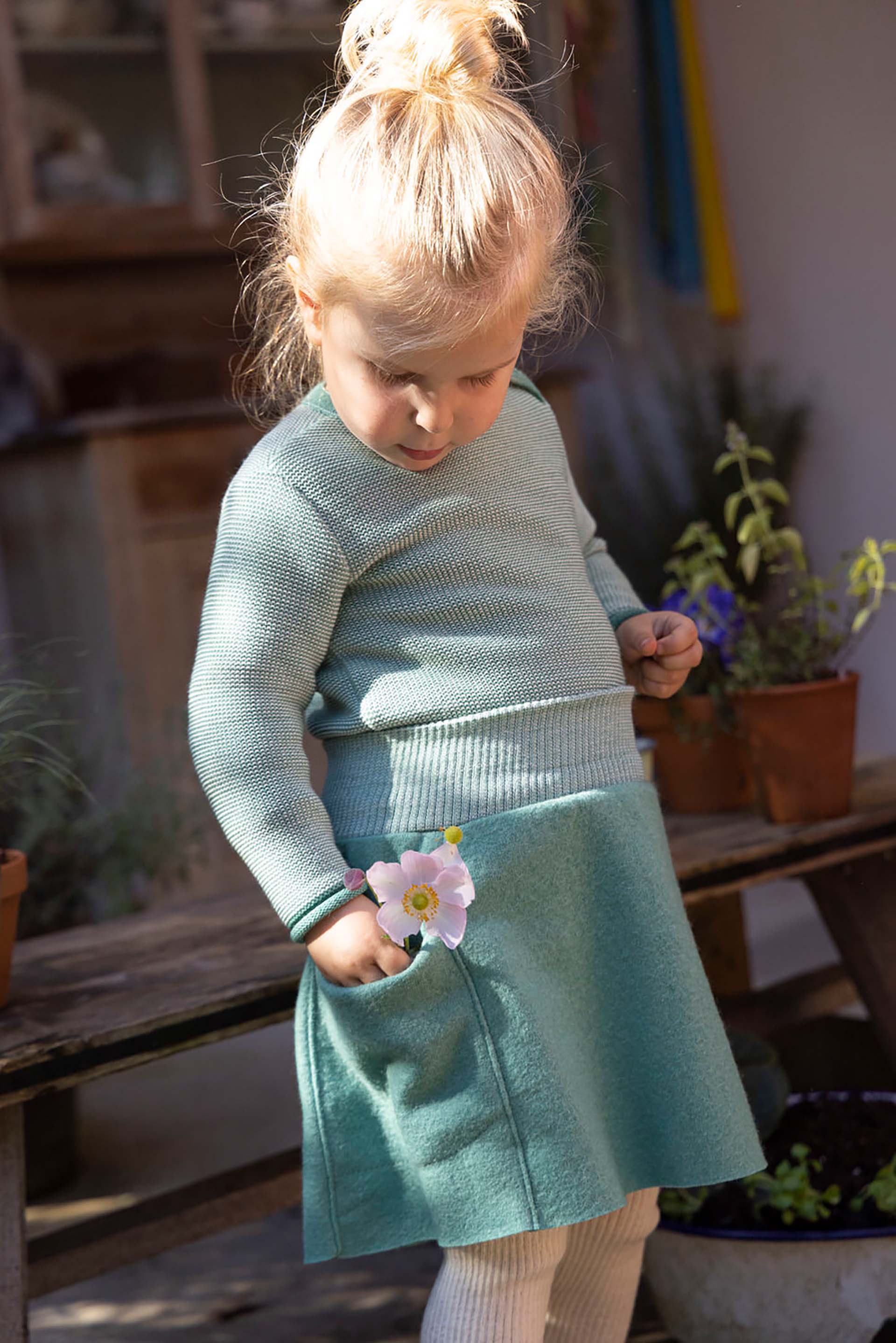 Blondes Mädchen in disana Melange-Pullover Mint-Natur und Walk-Rock Mint steckt eine weiße Blume in die Rockentasche