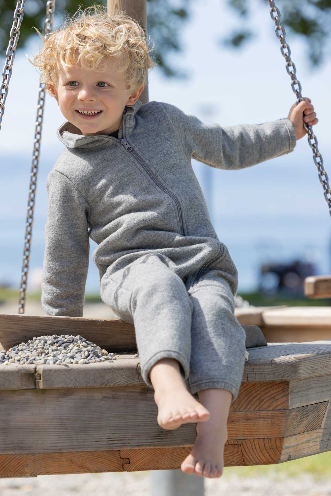 Blonder Junge im grauen disana Walk-Anzug aus leichtem Wollwalk aus Bio-Merinowolle sitzend auf einem Spielplatz-Holzpodest