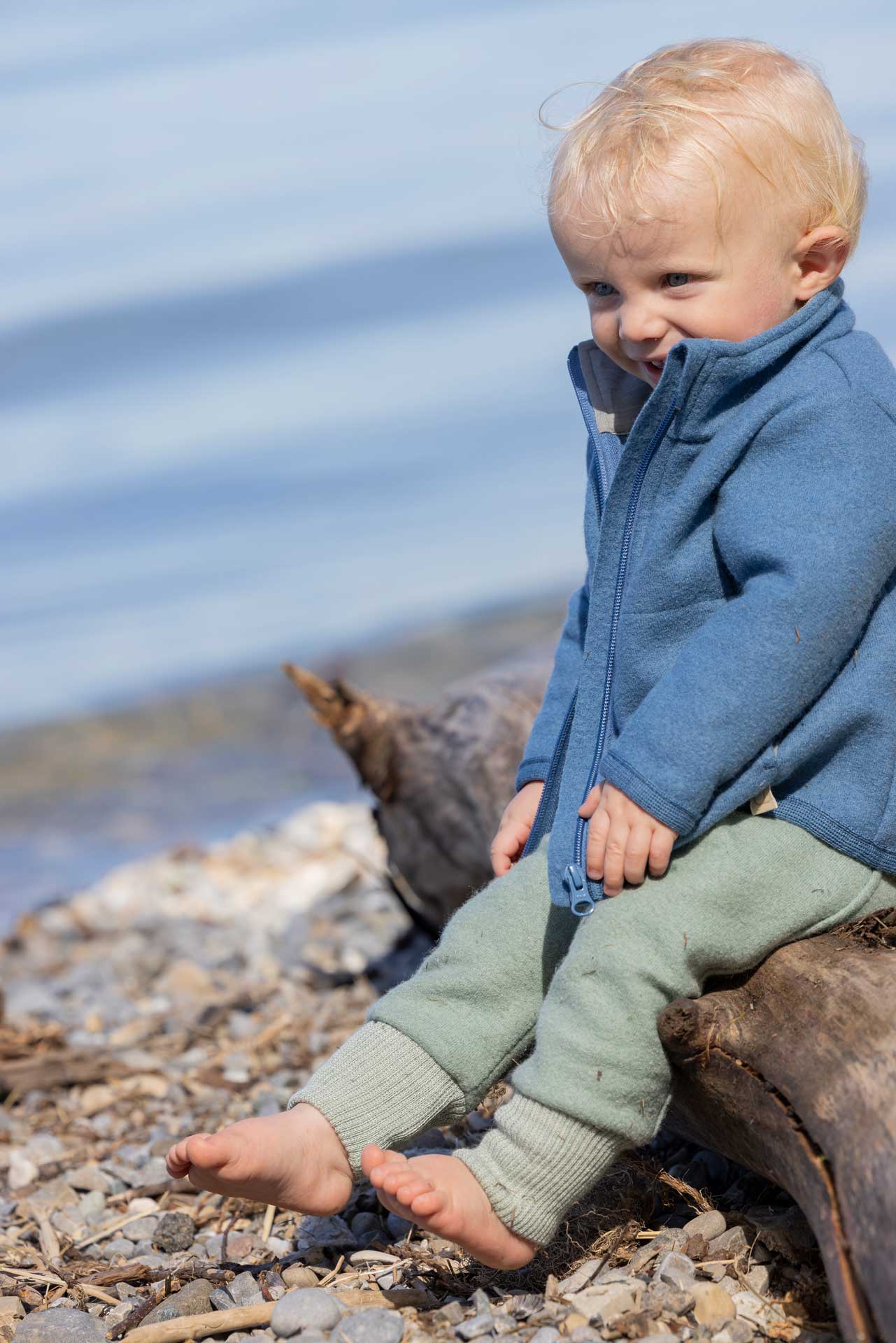 Blonder Junge in disana Zipper-Jacke Taubenblau & Pumphose Jade, beide aus leichtem Walk sitzend auf Treibholz am Kiesstrand