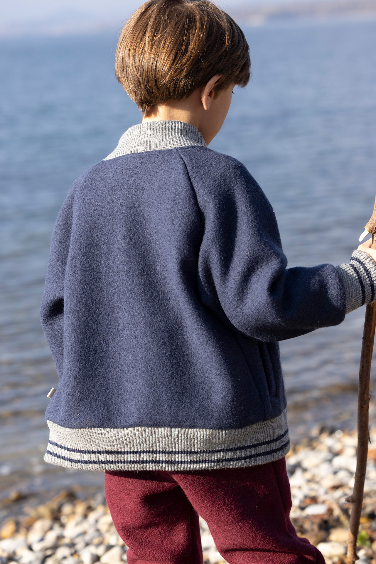 Rückansicht: Junge, braune Haare in disana College-Jacke Indigo mit grauen Strickbündchen, Walkhose in Cassis hält 1 Stock
