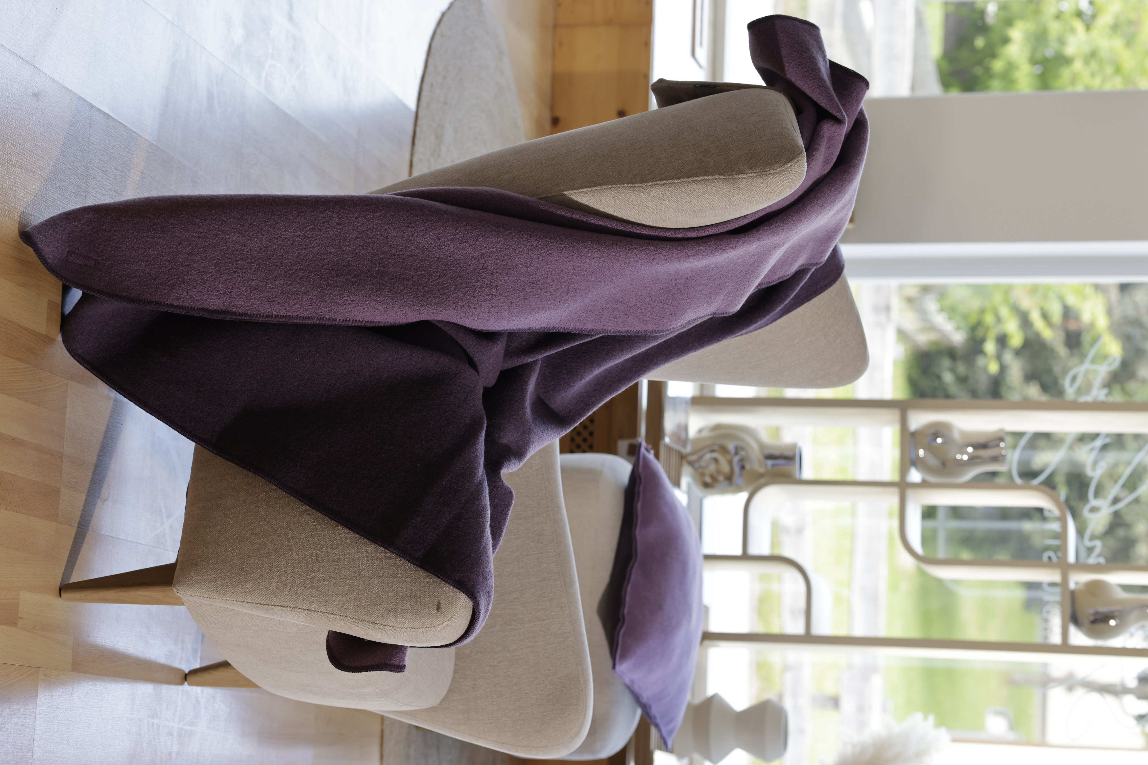 disana Doubleface-Decke in Aubergine-Flieder aus Bio-Merinowolle auf einem hellen Sessel in einem modern Wohnzimmer