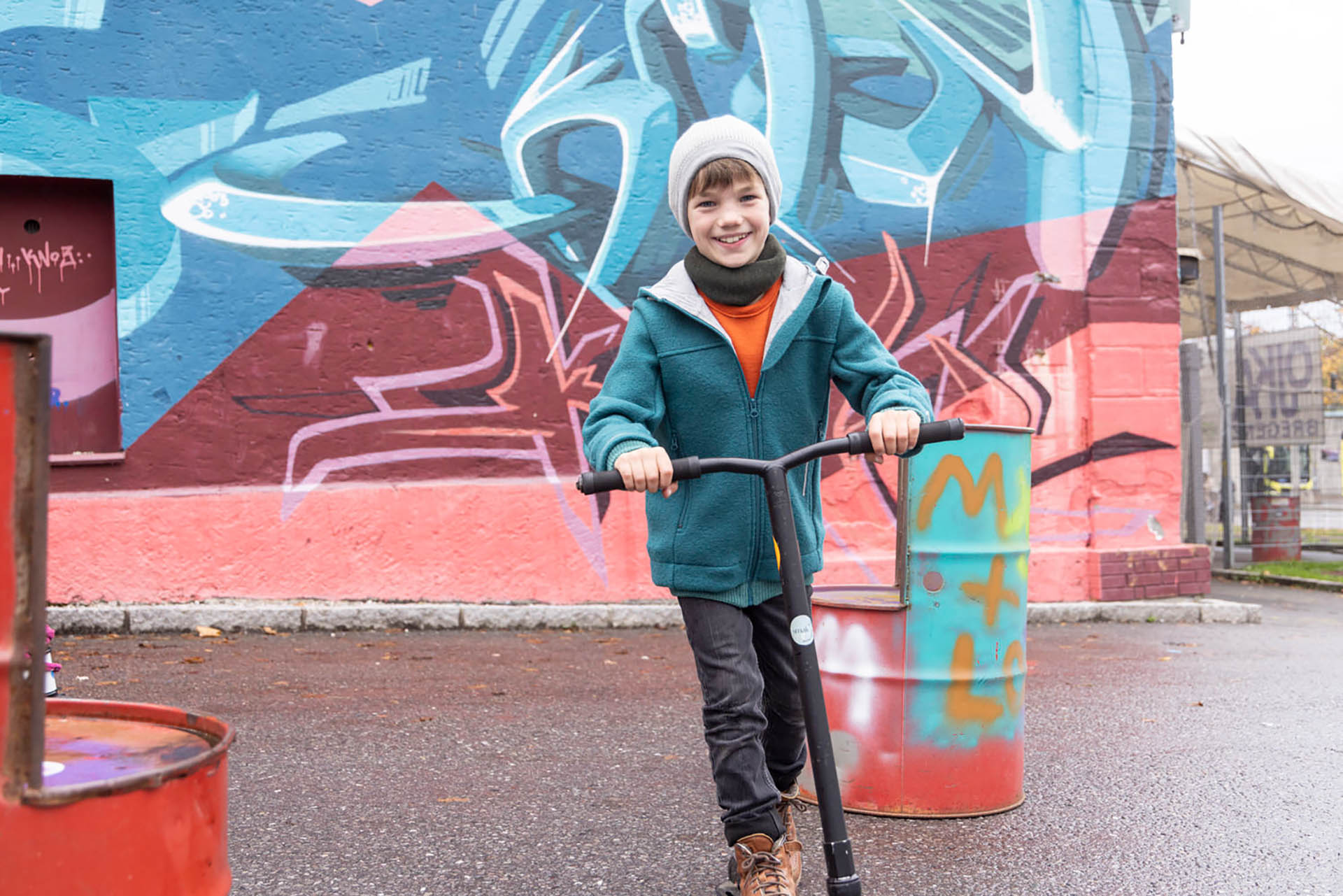 Junge in disana Outdoor-Jacke in Pacific und Beanie in Grau-Natur lacht auf einem Roller vor einer bunten Graffiti-Wand.