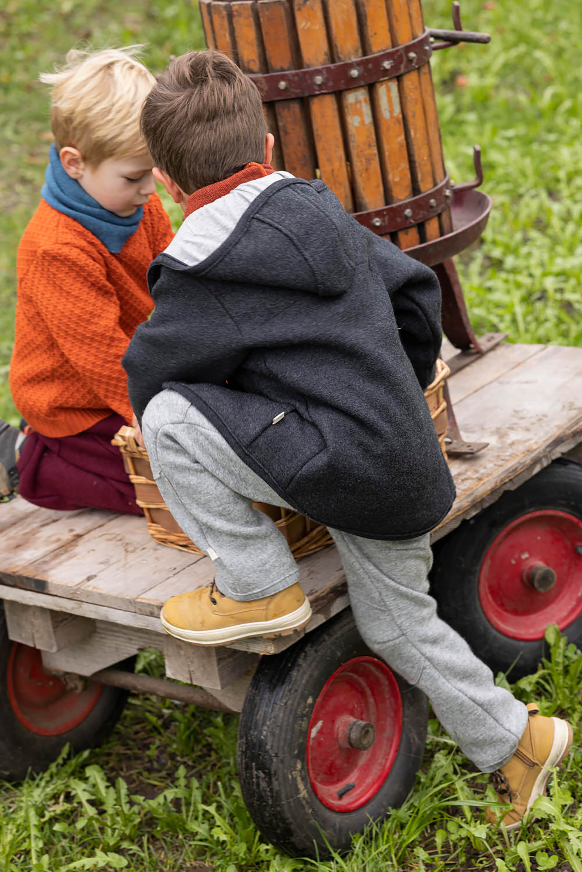 Zwei Jungen in disana Wollwalk-Kleidung spielen auf einem Holzwagen mit einer alten Obstpresse und Weidenkörben im Garten.